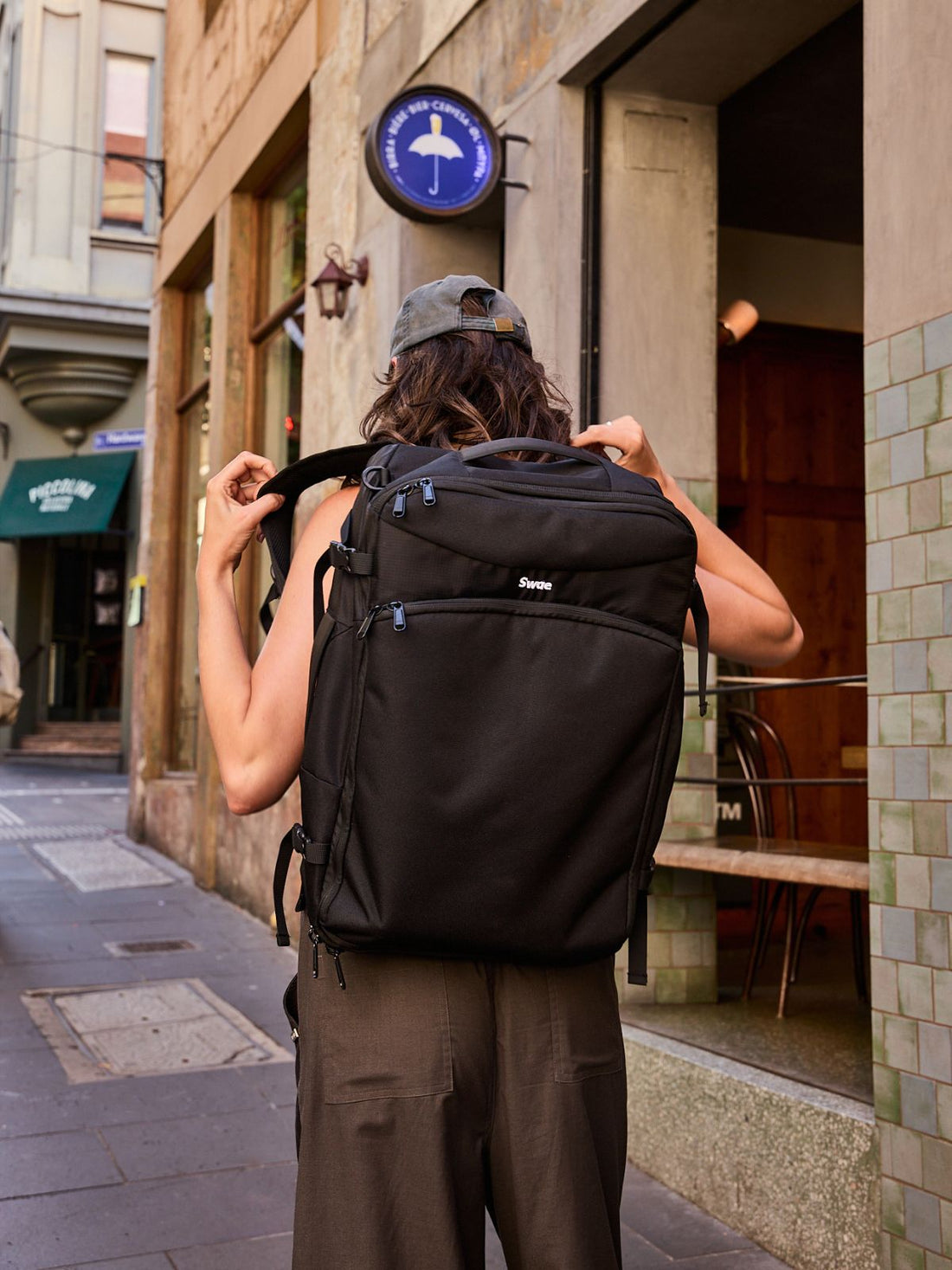 Man holding a black Swae Flow 40L Travel Backpack by the top handle, standing against a blue background in casual white outfit and sneakers.